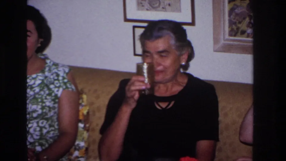 Women sit on a couch in a private living room, including an older woman in a black top holding a tall patterned glass.