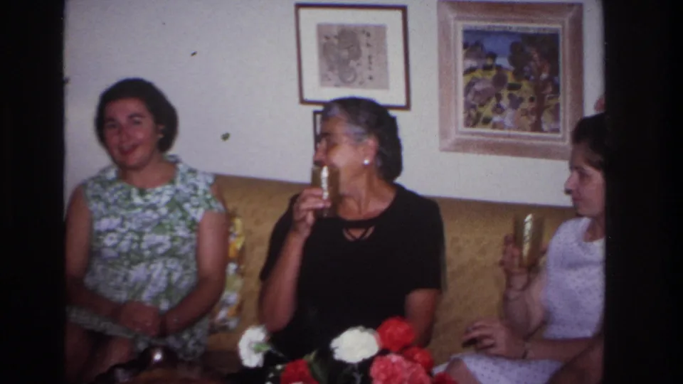 Three women in a living room sit on a couch with patterned glasses, framed pictures behind them, and flowers on a low table.