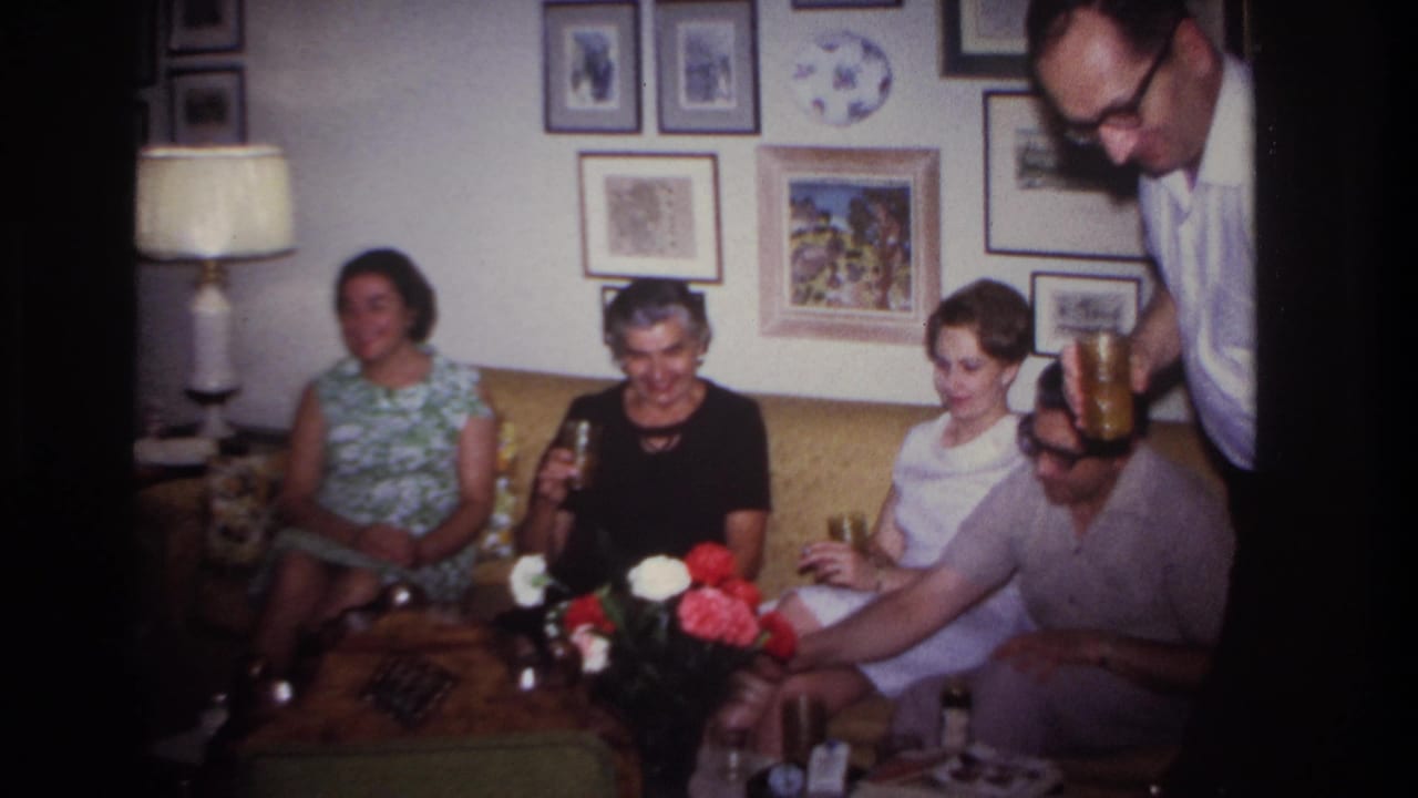 1967 Rockaway Beach home-movie frame showing adults gathered on a sofa in a private living room with drinks and mid-century decor.