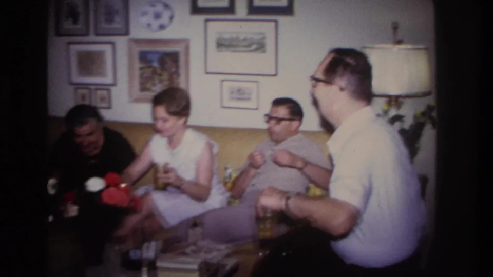 1967 Rockaway Beach home-movie frame showing adults in conversation inside a private living room with drinks and framed pictures.