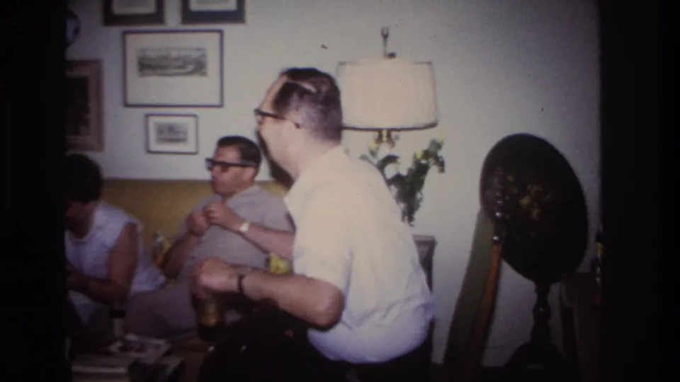 1967 Rockaway Beach home-movie frame with seated adults in a private living room, including two men in the foreground and mid-century furnishings.