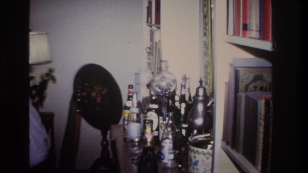 1967 Rockaway Beach home-movie frame showing a private living room with bottles, glassware, framed decor, and a partially visible adult.