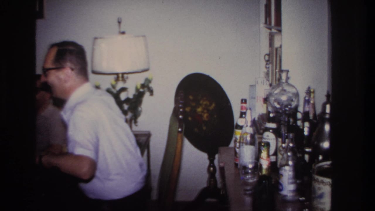 Man in glasses leans forward in a living room near a lamp and bottles.