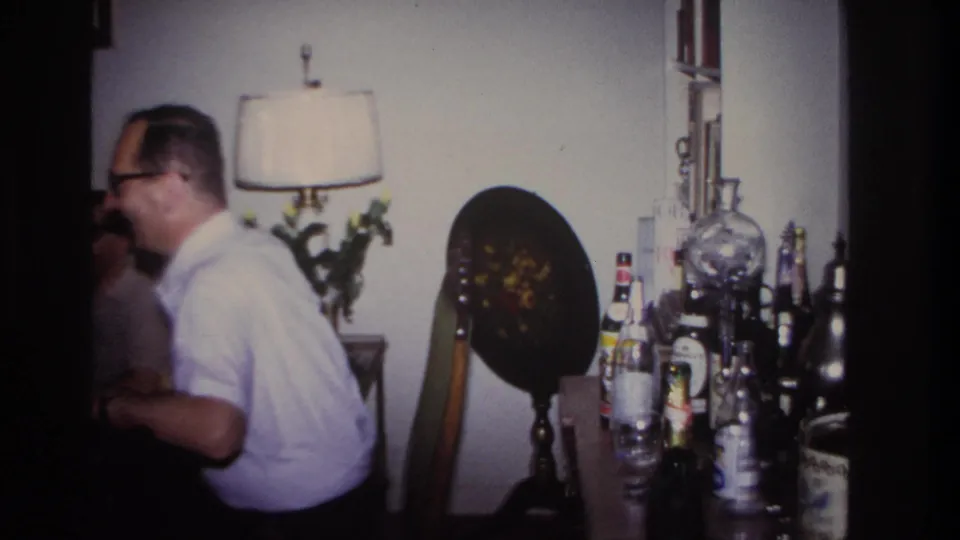 A man in glasses appears mid-conversation in the supplied 1967 Rockaway Beach living-room gathering footage.