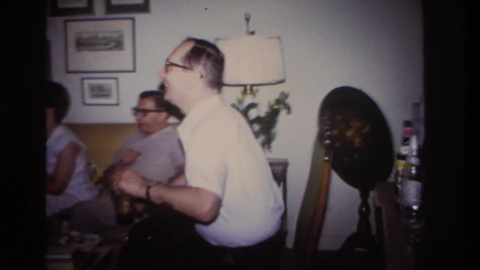 In the 1967 Rockaway Beach home-movie scene, a laughing man in glasses stands near seated adults in a decorated living room.