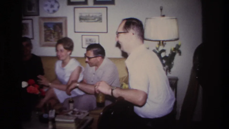 Adults sit together on a couch in a private Rockaway Beach living room as a man in glasses stands at the edge of the frame.