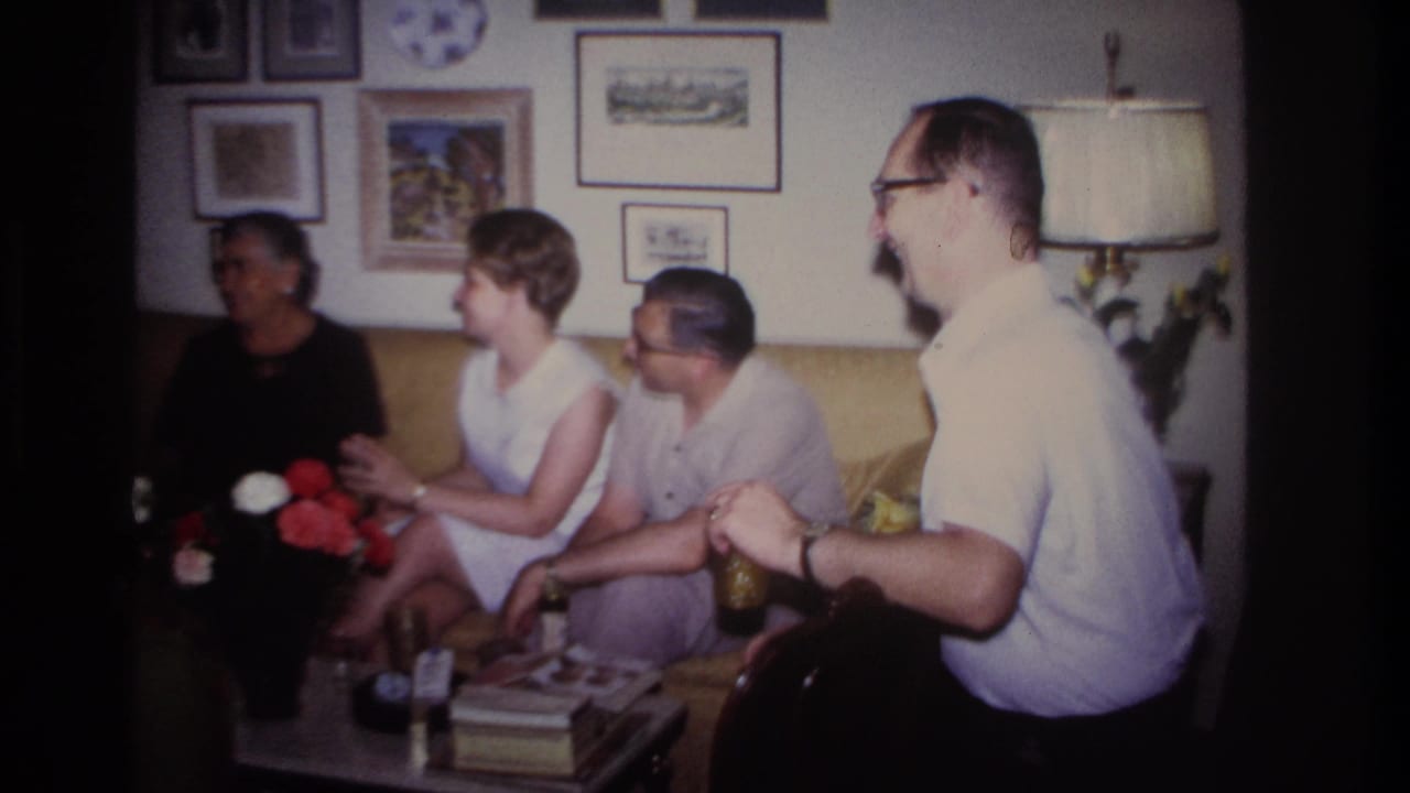 1967 Rockaway Beach home-movie frame showing adults gathered on a yellow sofa in a decorated living room.