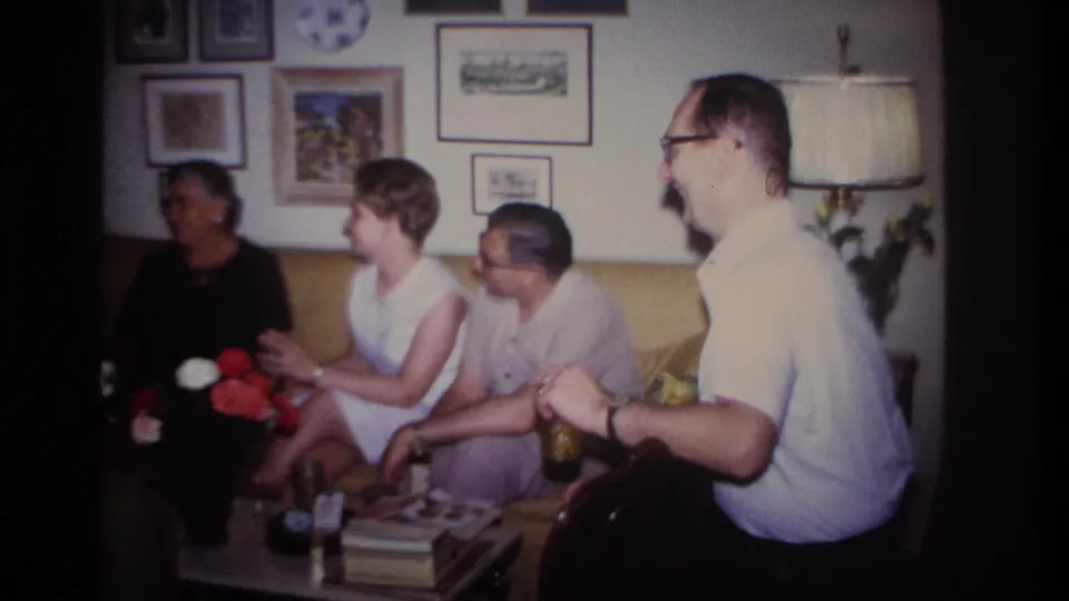 1967 Rockaway Beach home-movie frame showing adults gathered on a yellow sofa in a decorated living room.