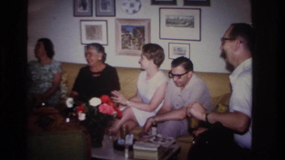 Adults converse and smile in a 1967 Rockaway Beach living room, with flowers and framed wall art visible.