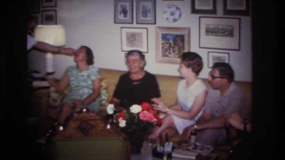 1967 home-movie frame of adults laughing in a Rockaway Beach living room as a person reaches in from the side.