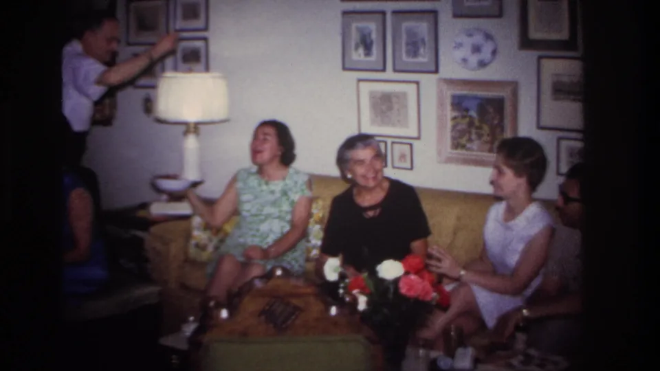 Rockaway Beach 1967 home-movie frame showing adults on a sofa in a private living room with lamp, flowers, and wall art.