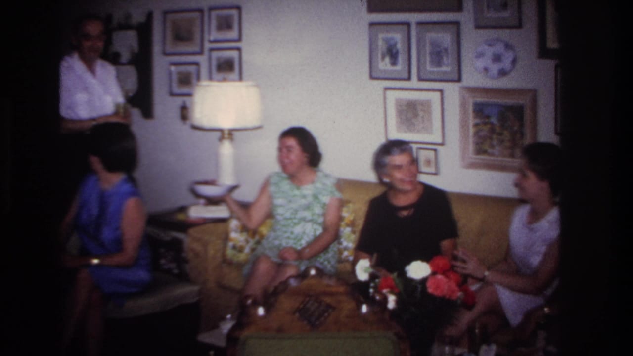 1967 Rockaway Beach home-movie frame of adults seated in a living room while a man stands near the wall and others talk on a sofa.