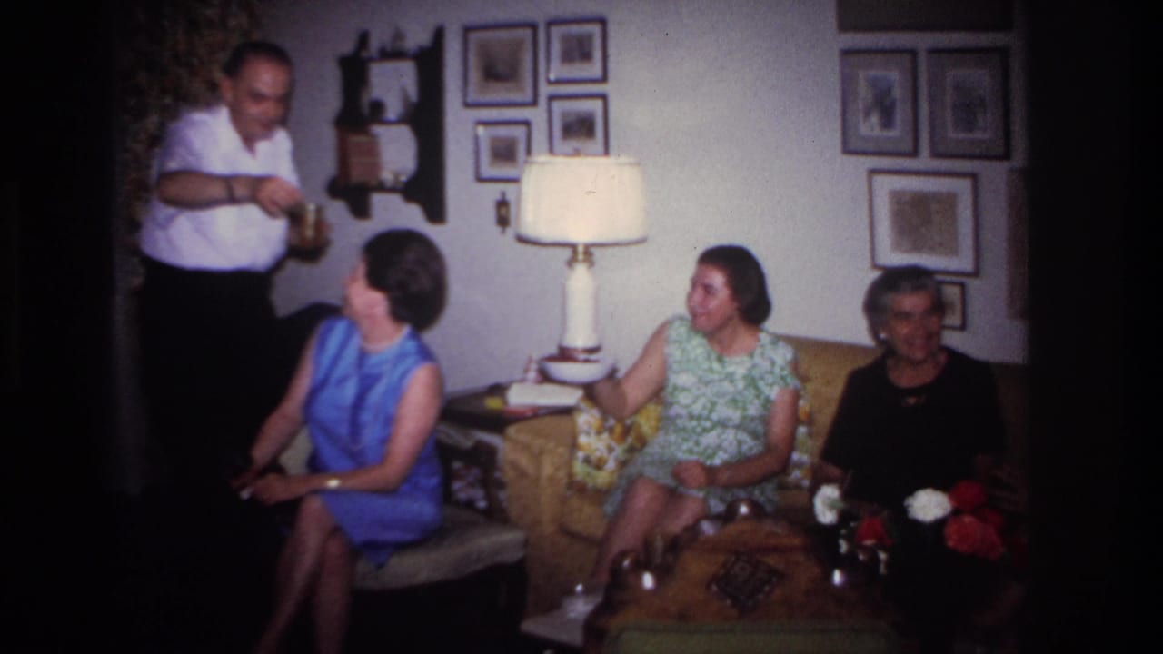 Man stands holding food or drink while women sit near a sofa and lamp.