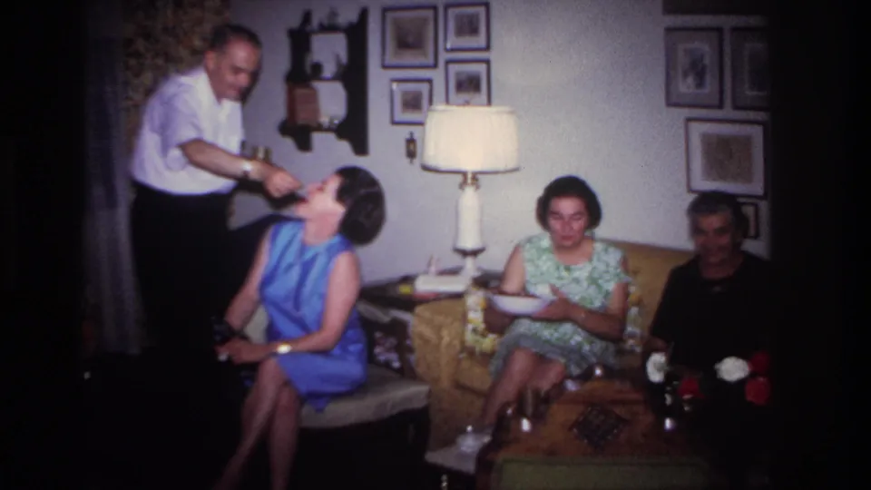 1967 Rockaway Beach home-movie frame of a man offering food to a seated woman in a blue dress during a living-room party.