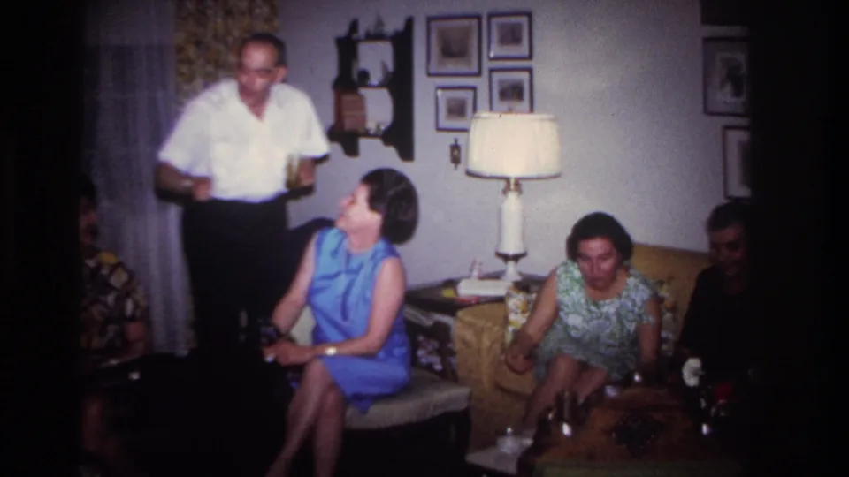 1967 Rockaway Beach interior with a man holding a glass beside seated adults, a lamp, wall pictures, and flowers on the table.