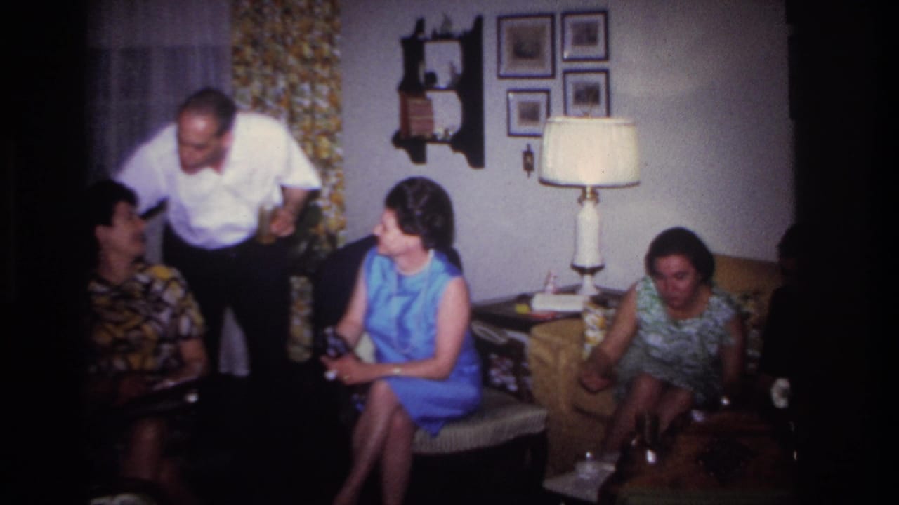 1967 Rockaway Beach home-movie frame showing adults conversing in a private living room with floral curtains and a table lamp.