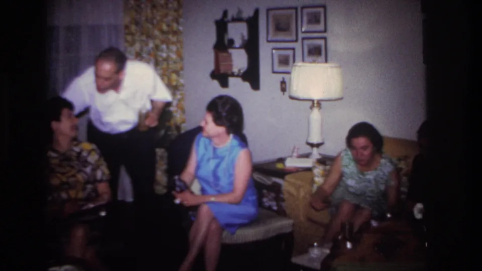 1967 Rockaway Beach home-movie frame showing adults conversing in a private living room with floral curtains and a table lamp.