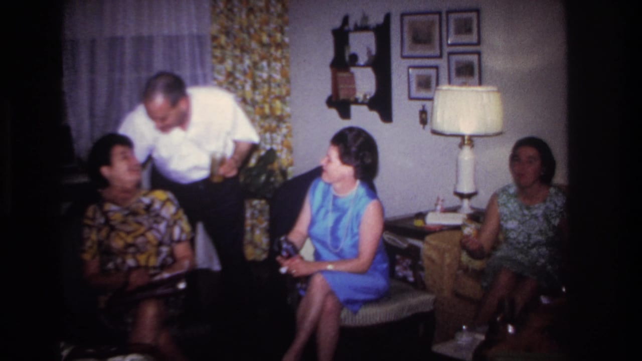 A man stands beside three seated women during an indoor family conversation.