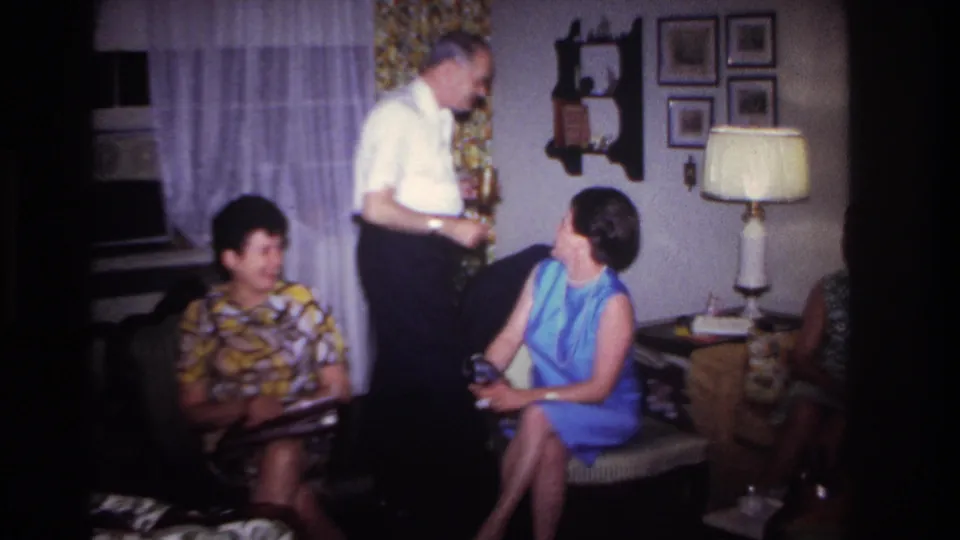 1967 home-movie frame from Rockaway Beach showing a man standing near a woman in a blue dress and another seated woman.