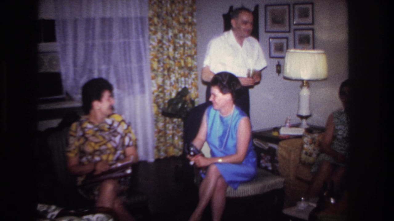 Adults in a living room as a standing man holds a drink and seated women look toward each other.
