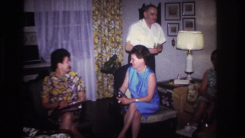 1967 Rockaway Beach home-movie frame of adults in a private living room, with a standing man holding a drink and seated women conversing.