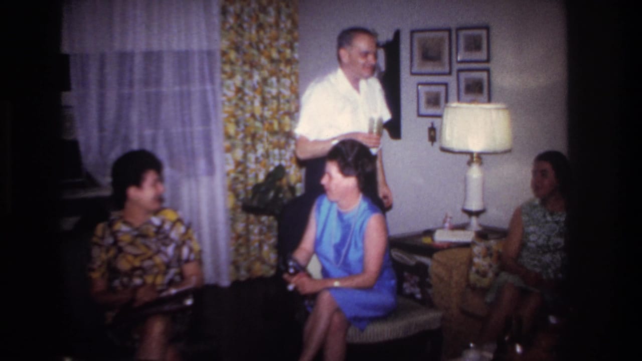Adults gather in a Rockaway Beach living room in 1967 as a man in a white shirt holds a drink near seated women.