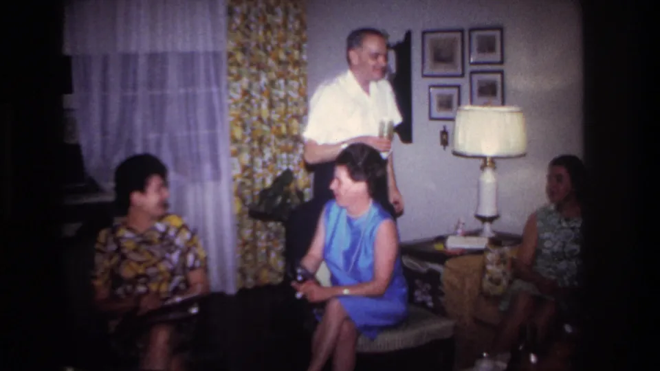 Adults gather in a Rockaway Beach living room in 1967 as a man in a white shirt holds a drink near seated women.