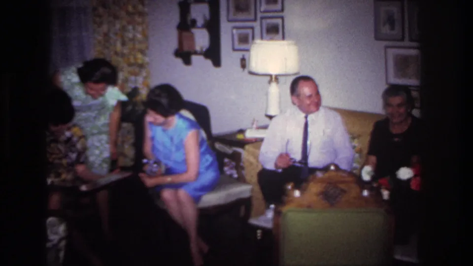 1967 home-movie frame from Rockaway Beach showing women looking at a photo album as other adults sit nearby.