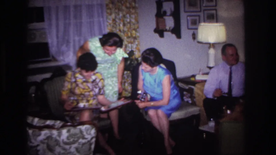 Women in patterned and blue dresses study a photo album in a 1967 Rockaway Beach living room as a man sits to the side.