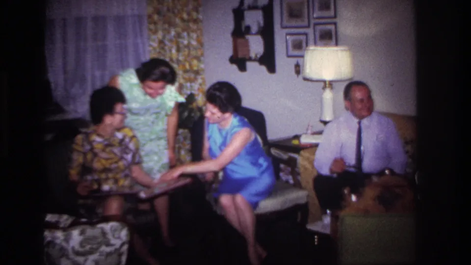 1967 Rockaway Beach home-movie frame of adults in a living room as a woman in a blue dress shows photographs to seated relatives.