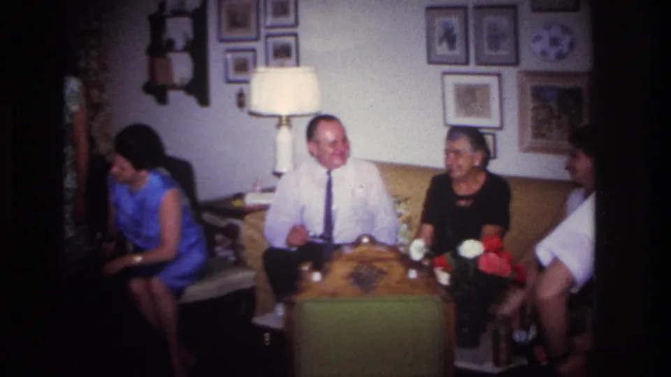 1967 Rockaway Beach home-movie still showing adults seated around a coffee table in a private living room with flowers and wall art.