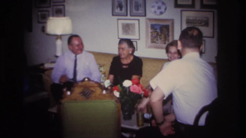 Home-movie frame from the 1967 Rockaway Beach gathering, showing adults talking around a coffee table in a private living room.