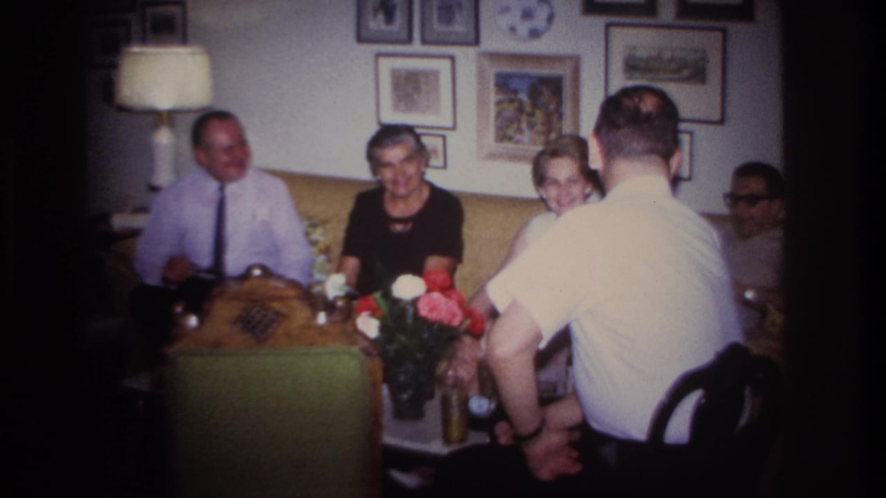 Four adults seated around a coffee table in a living room with framed pictures and a lamp.