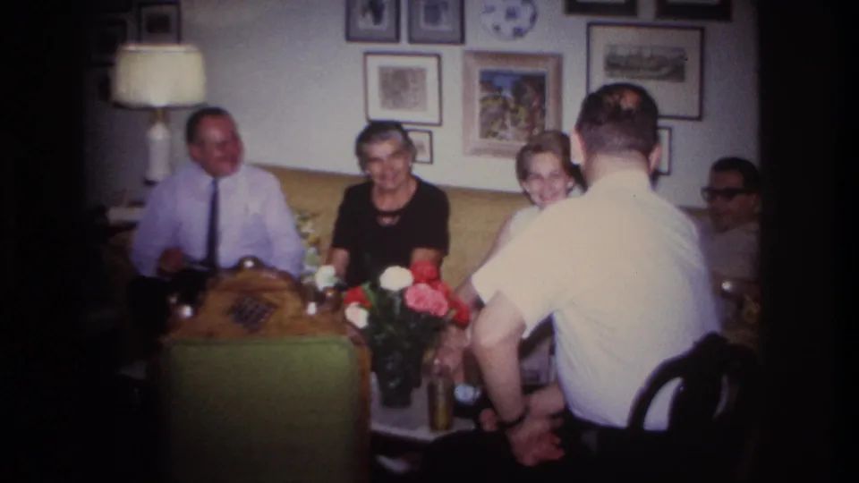 1967 Rockaway Beach home-movie frame showing adults gathered in a living room with a yellow sofa, flowers, framed wall art, and table lamp.