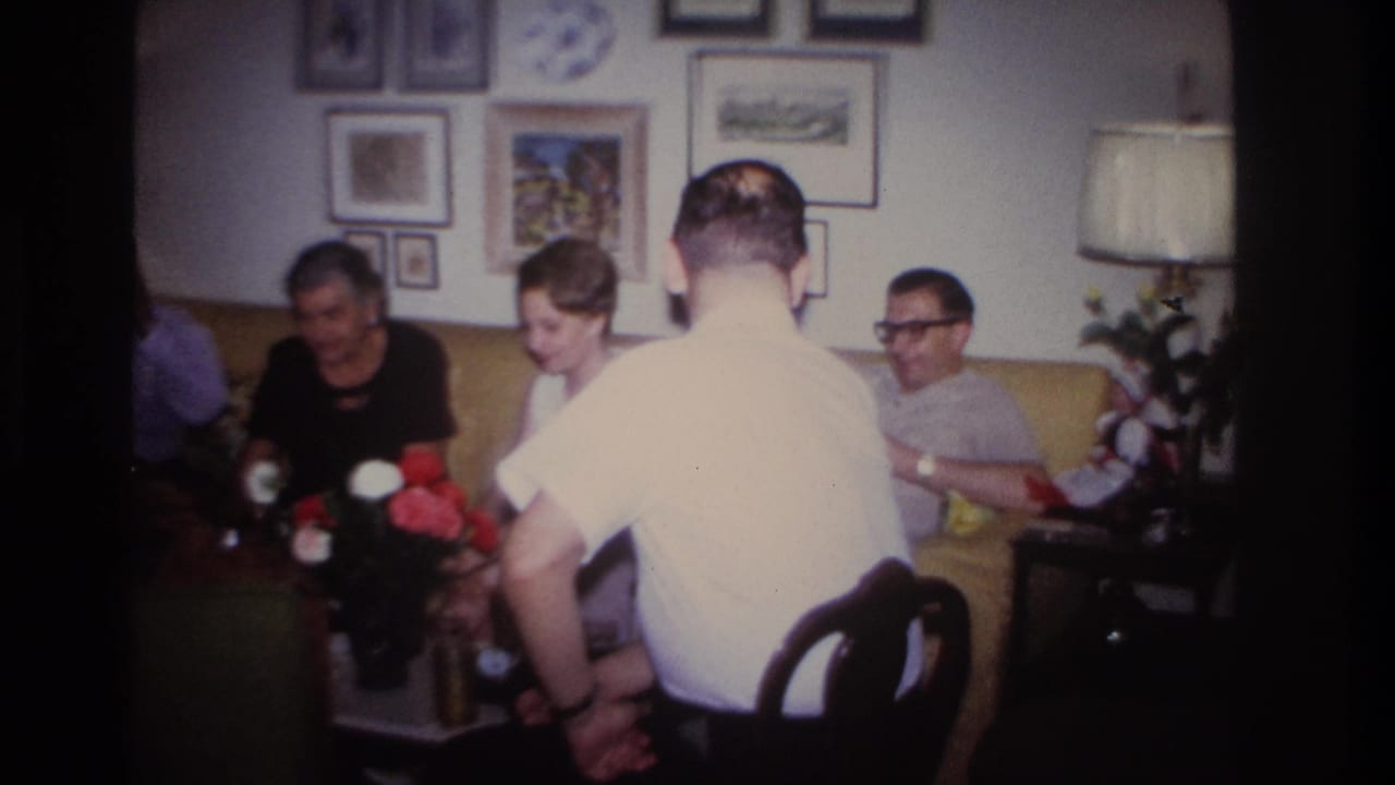 Adults sit on a yellow couch while a man in a white shirt faces away from the camera.