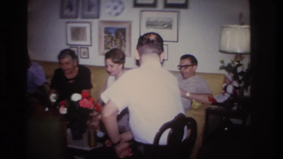 1967 Rockaway Beach interior frame with adults seated on a yellow couch as a man in a white short-sleeve shirt faces the group.