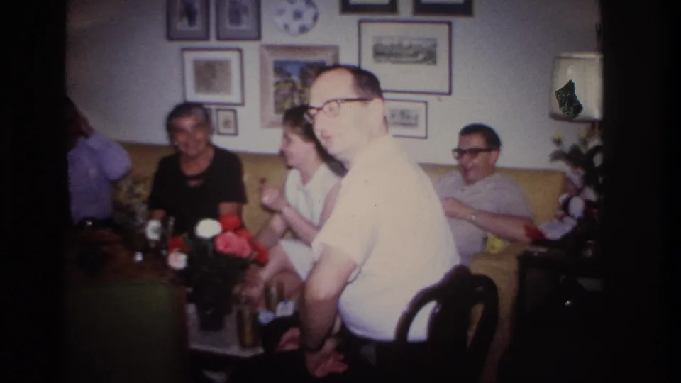 1967 Rockaway Beach home-movie frame showing a bespectacled man turning toward the camera during a living-room conversation.