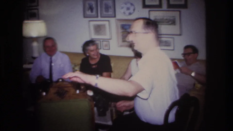 1967 Rockaway Beach living-room frame with a man in a white shirt gesturing near a coffee table while adults sit behind him on a yellow sofa.