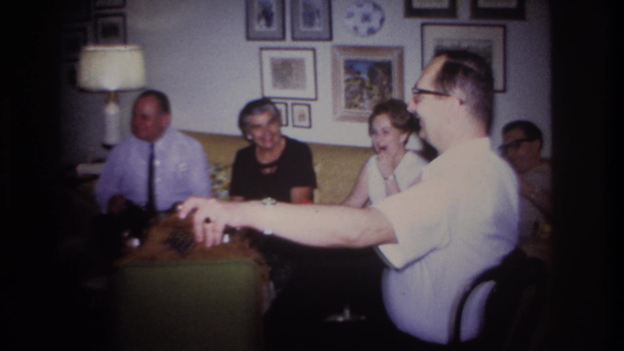 Adults sit in a living room while a man in glasses gestures from a chair near a table.
