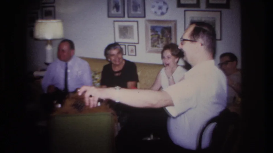 1967 Rockaway Beach home-movie frame of adults gathered on a yellow couch as a man in glasses gestures in a private living room.