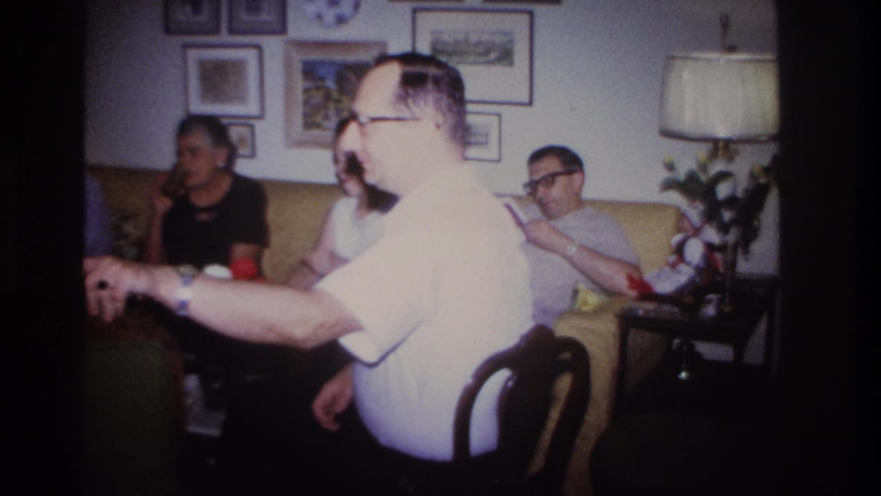 A man in a white shirt gestures beside adults seated on a couch in a living room.