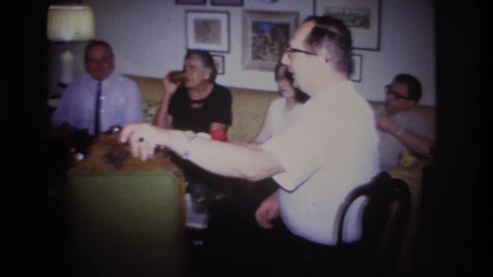 Adults in a private Rockaway Beach living room watch and laugh as a man in glasses gestures near the camera.