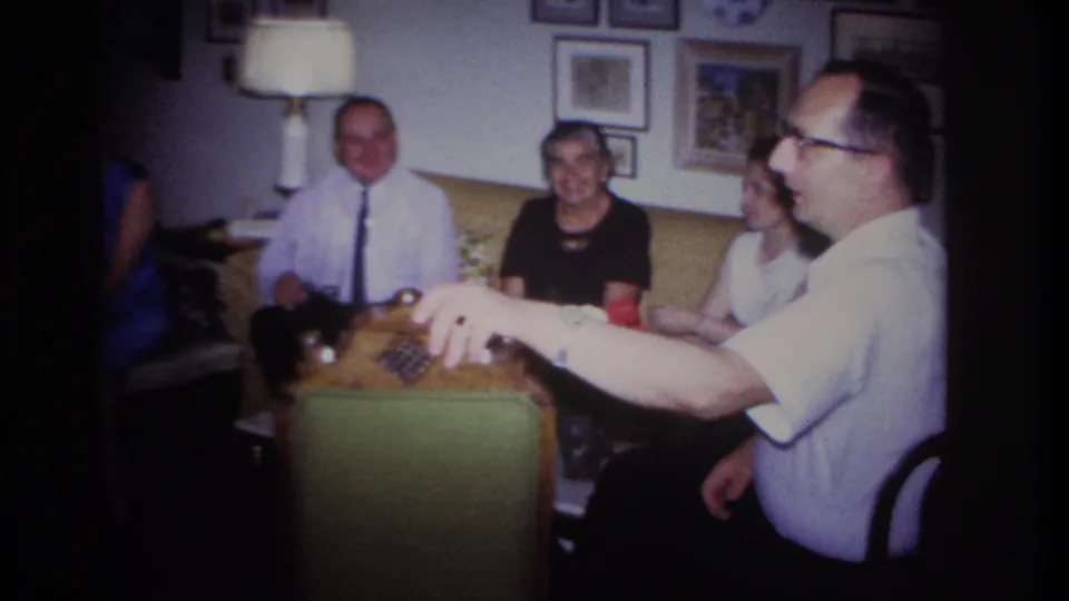 1967 Rockaway Beach home-movie frame of adults seated in a private living room while a man gestures from the right foreground.