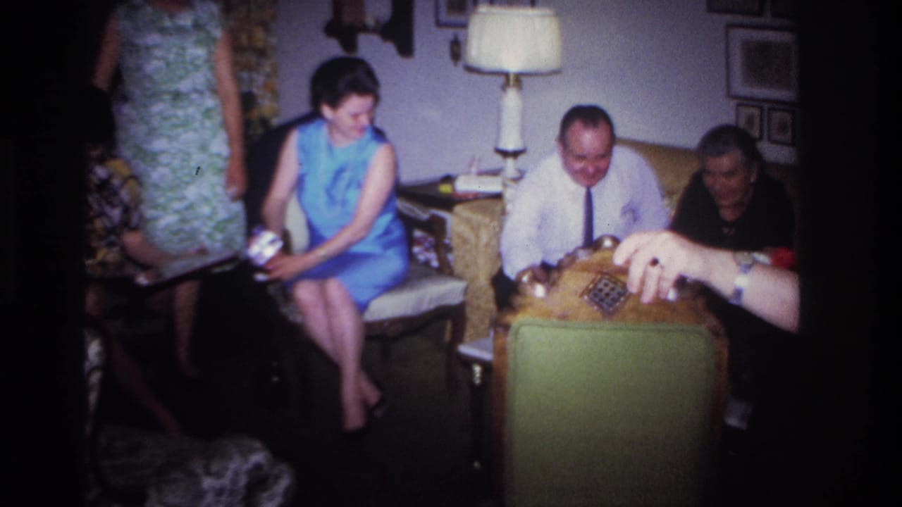 Adults seated in a living room with a lamp, framed wall art and a chair in the foreground.