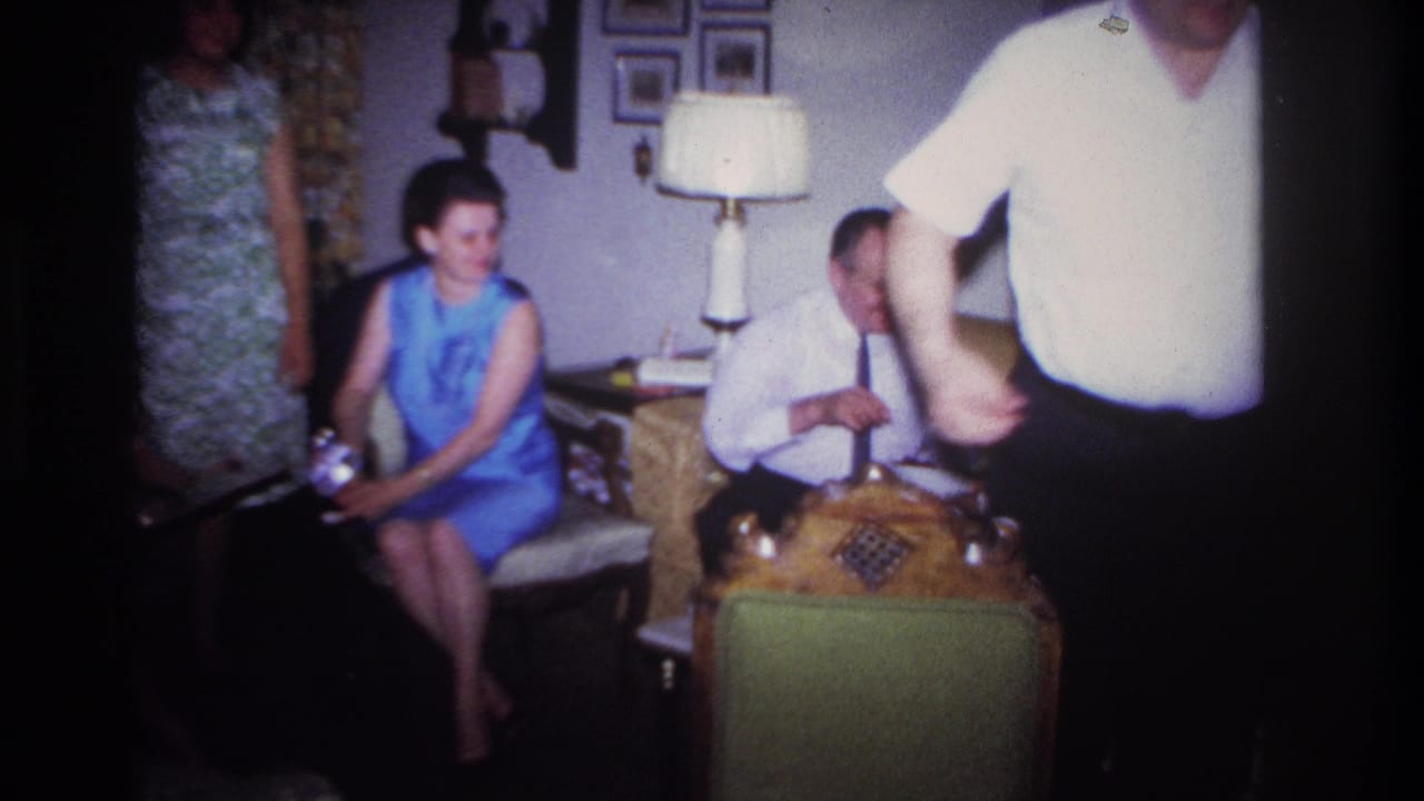 In the Rockaway Beach living room, a woman in a blue dress sits beside a man in a white shirt and tie as someone moves through the foreground.