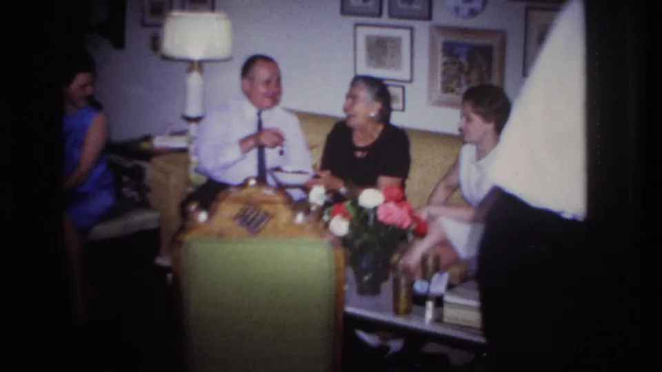 Adults converse in a 1967 Rockaway Beach living room, with flowers on a table, framed pictures behind them and a person partly in the foreground.