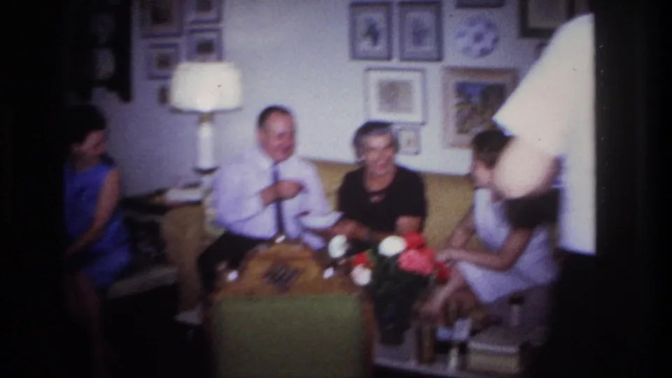 A group of adults smiles and talks in a Rockaway Beach living room, with a table lamp, framed pictures, flowers and a foreground figure visible.
