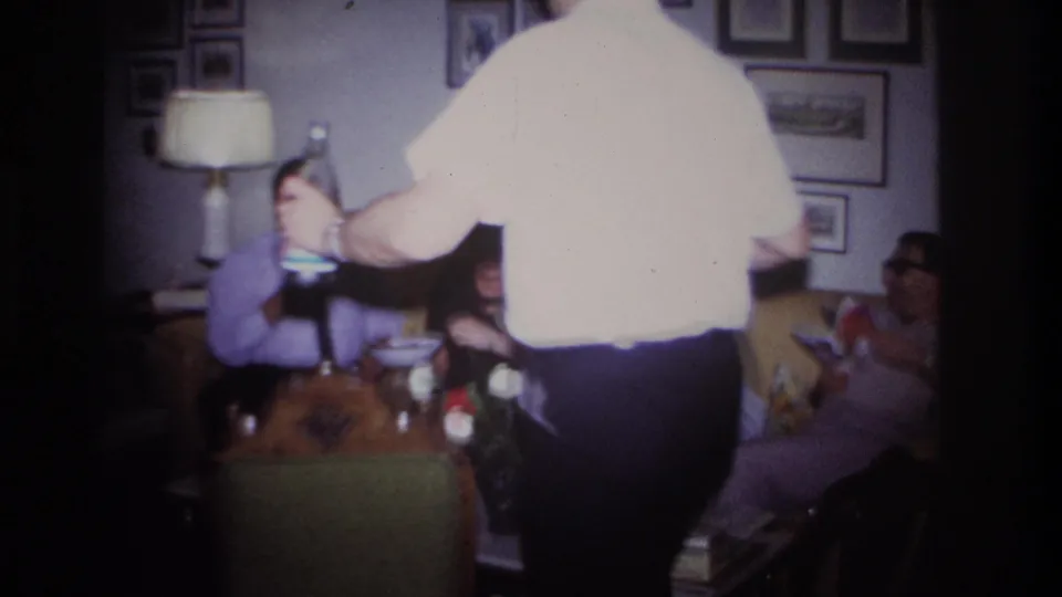 1967 Rockaway Beach home-movie frame of adults on a yellow couch as a man in a white shirt serves drinks in a framed-wall living room.