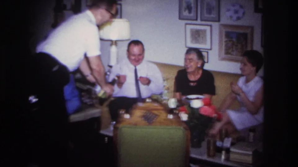 A handheld 8mm frame shows a man leaning in to serve drinks to adults seated on a yellow couch in a 1960s living room.