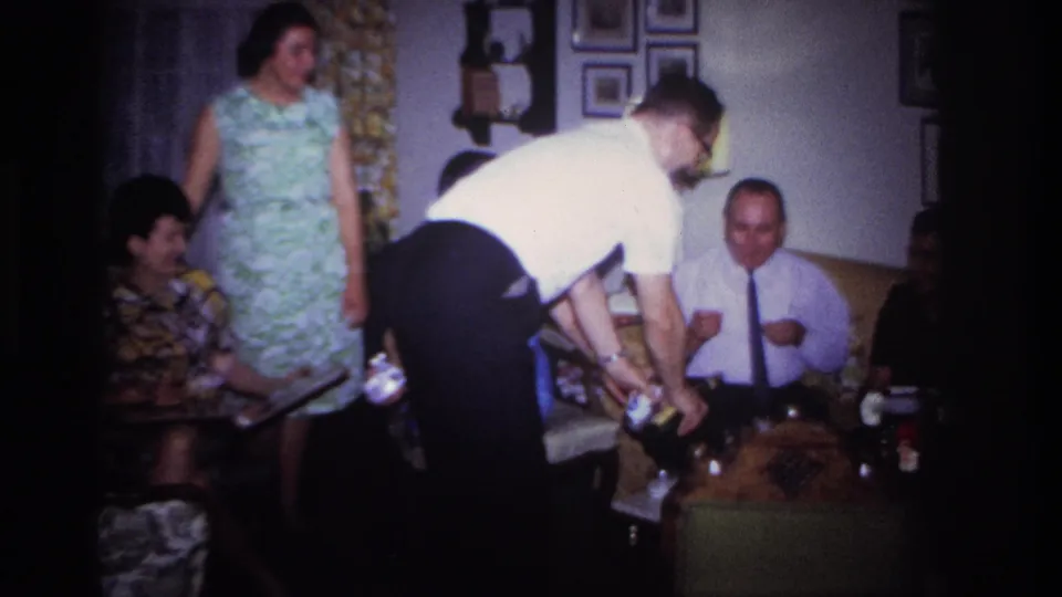 In the Rockaway Beach living room, a woman in a patterned dress stands as adults sit nearby and a man bends over the table with bottles.